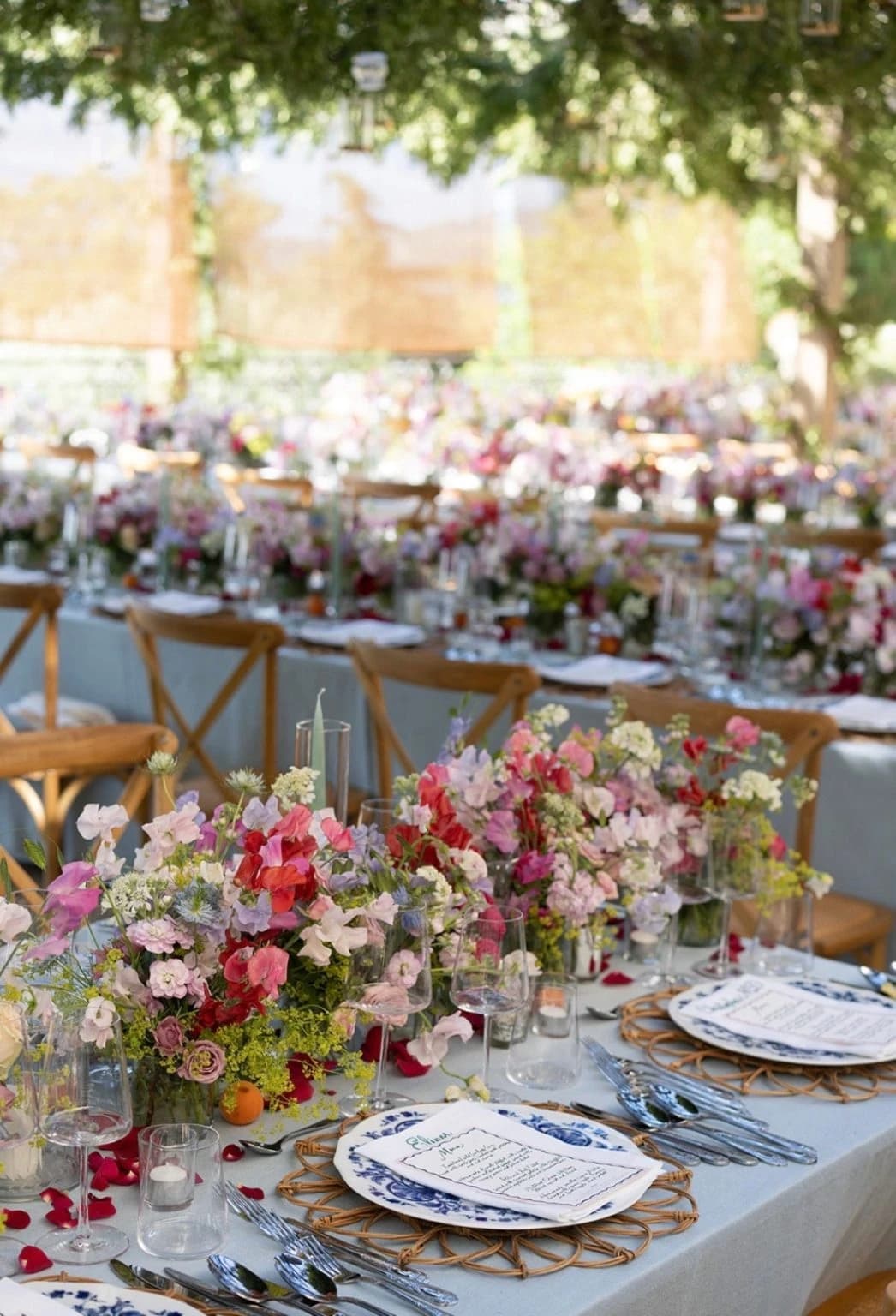 Une longue table dressée sous les arbres : nappe bleu pâle, sets en rotin, menus manuscrits et fleurs des champs sur toute la longueur.