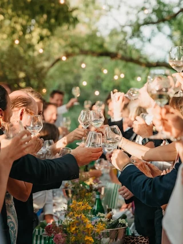 Des invités lèvent leurs verres au-dessus d’une longue table, sous des guirlandes lumineuses, à la tombée du jour.
